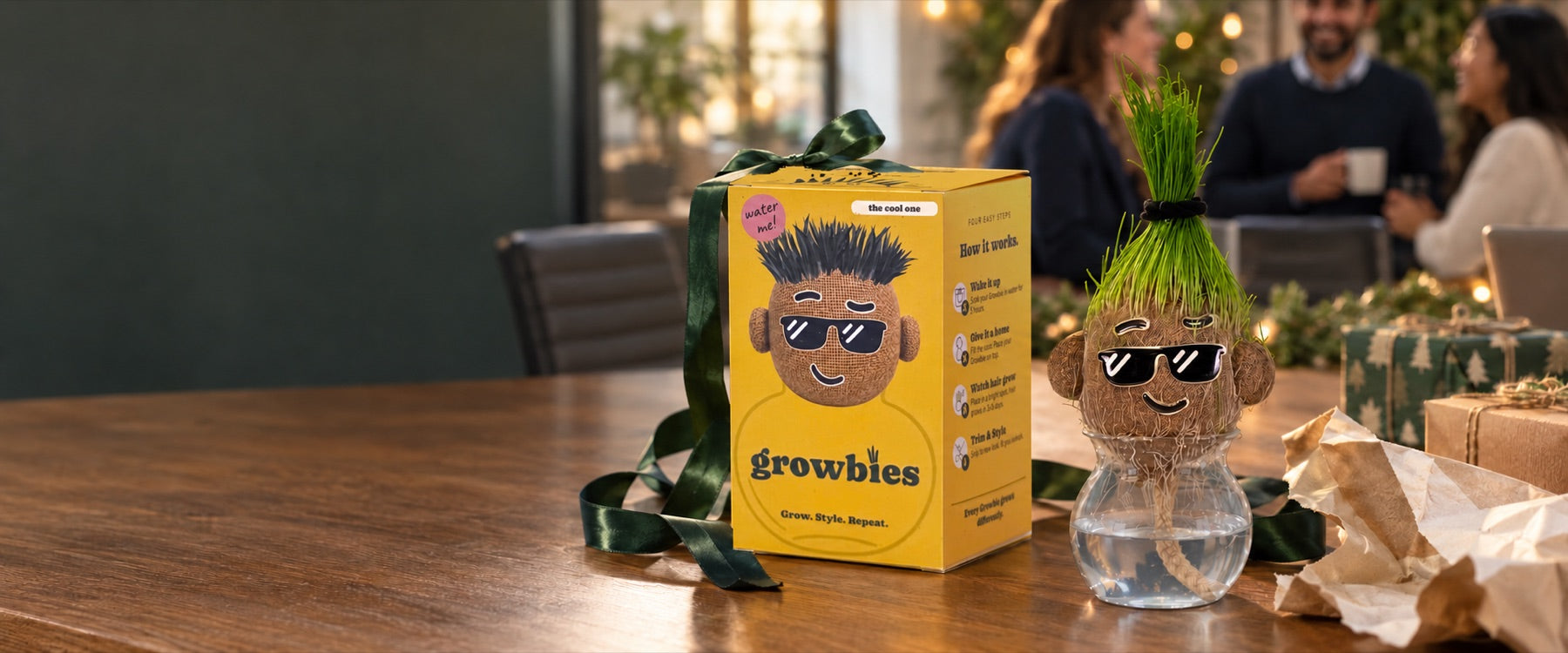 A boxed Growbie with a green ribbon next to a grown one with grass hair, on the table at an office gift exchange